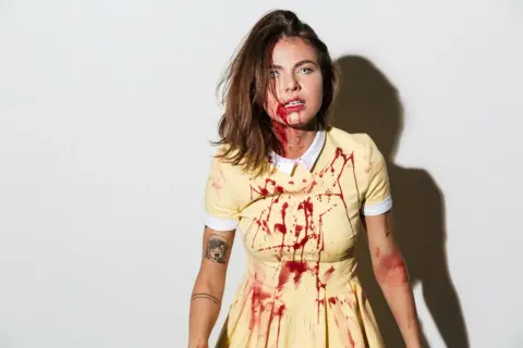 Przerażający strój na Halloween: dziewczyna w żółtej sukience z plamami krwi, z makijażem sugerującym ranę.