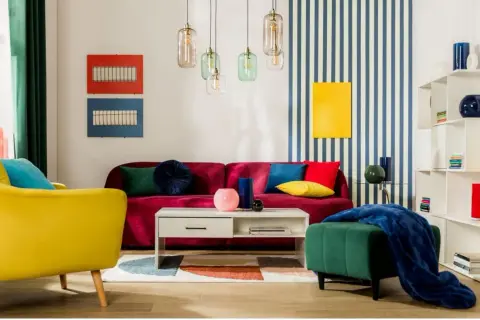 Salon w stylu memphis: żółty fotel, bordowa kanapa, kolorowe poduszki, geometryczny dywan i lampy.