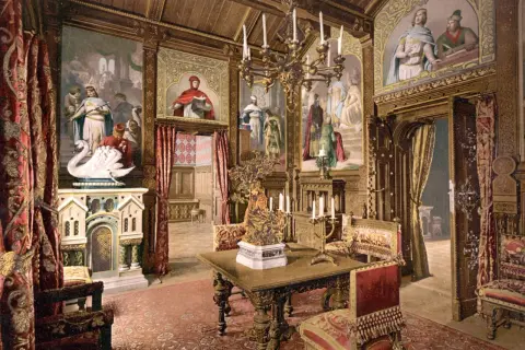 Prunkvoller Salon im Schloss Neuschwanstein innen. Wandmalereien mit Sagenmotiven, ein Kamin mit Schwan, prunkvolle Möbel und ein Kronleuchter schaffen königliches Ambiente.