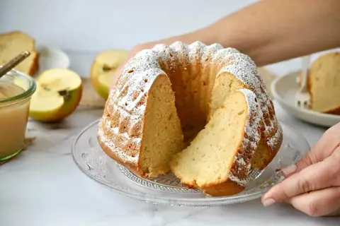 Saftiger Gugelhupf mit Puderzucker bestäubt, daneben Äpfel und Apfelmus. Ein Stück ist bereits angeschnitten.