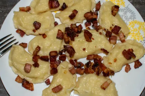 Pyszne pierogi galicyjskie posypane skwarkami z boczku, gotowe do zjedzenia. Obok leży widelec.