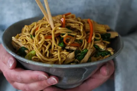 En skål med lo mein-nudlar, kyckling och grönsaker, lyfts med ätpinnar.