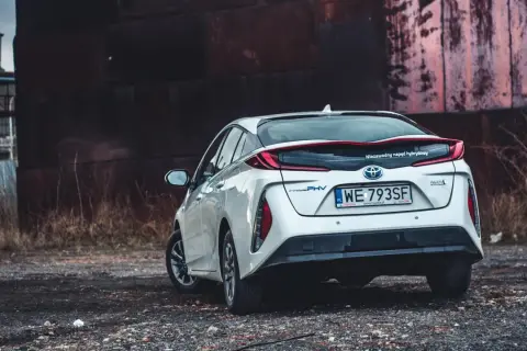 Biały Prius 4 z napędem hybrydowym stoi na tle industrialnego krajobrazu.
