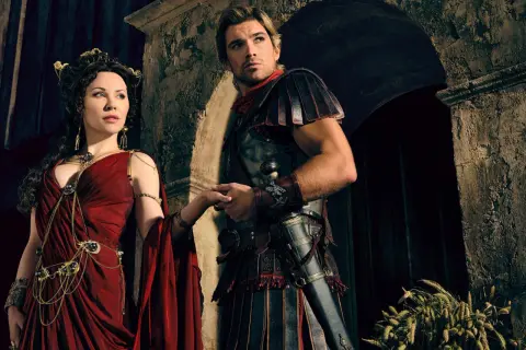 Die Besetzung von Spartacus: Ashur und Lucretia, in prachtvollen Gewändern und Rüstungen, halten Händchen vor einer antiken Kulisse.