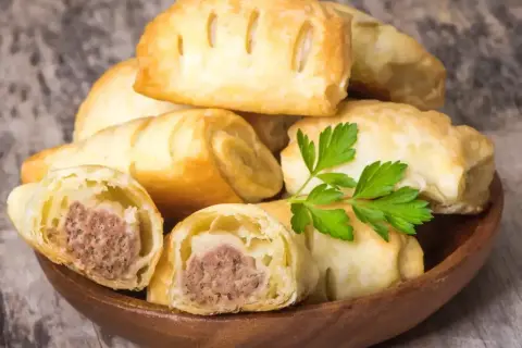Stos pieczonego ciasta francuskiego z kiełbaską w środku, ozdobione natką pietruszki.