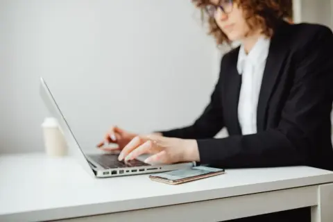 Kobieta w okularach i czarnej marynarce pracuje na laptopie. Obok leży telefon. Może szuka informacji, co to jest upadłość konsumencka.