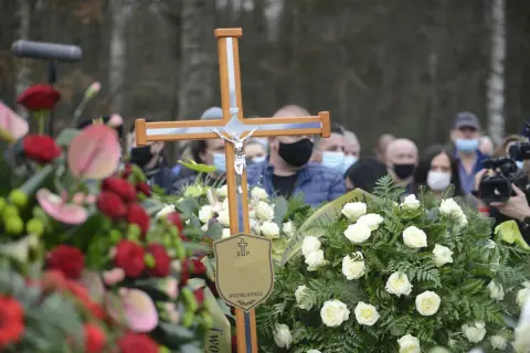 Drewniany krzyż z Jezusem, otoczony wieńcami, symbolizuje pożegnanie. Po pogrzebie tabliczka z napisem "Spoczywaj w pokoju" pozostaje jako pamiątka.