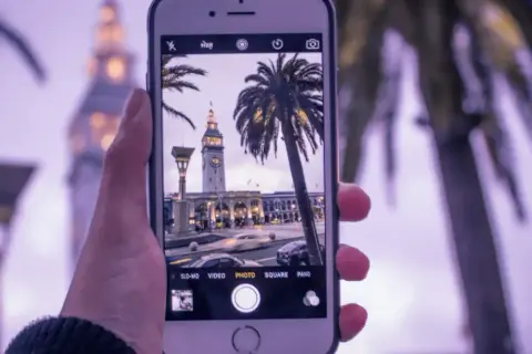 Dłoń trzyma iPhone'a, na ekranie widać budynek z zegarem i palmy. Dowiedz się, jak ustawić siatkę w aparacie iPhone, by ułatwić kompozycję.