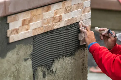 Close-up of a worker's hands carefully performing wall tile installation, applying a new stone tile to a textured adhesive layer.