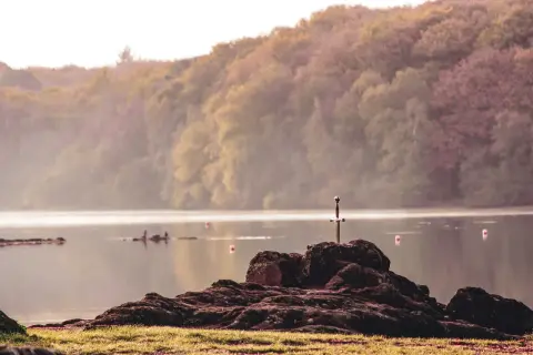 Épée plantée dans un rocher au bord du lac de Trémelin, entouré de brume et de collines boisées.