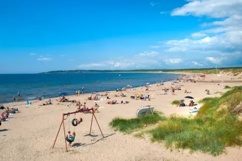 En solig strand i Halland fylld med badande och solande människor. Barn leker på en gunga nära sanddynerna.
