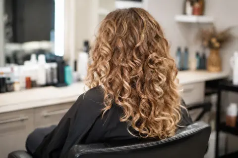 Kobieta w czarnym szlafroku, z pięknymi, złotymi loki trwała. W tle widać salon fryzjerski.