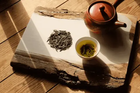 Théière en terre cuite, feuilles de bancha sèches et une tasse de thé vert infusé, sur un plateau en bois brut éclairé par le soleil.