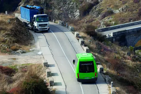 Camión blanco con contenedor azul subiendo una pendiente, seguido por un autobús verde. Se debate quién tiene prioridad en una pendiente.