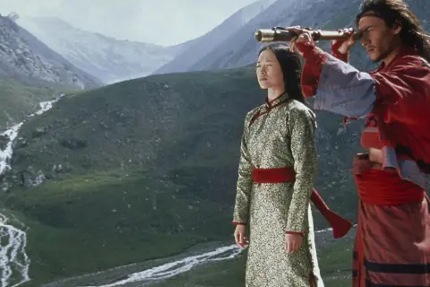 Para w tradycyjnych strojach, w tle górzysty krajobraz. Scena z kina azjatyckiego, gdzie mężczyzna przez lunetę obserwuje horyzont.