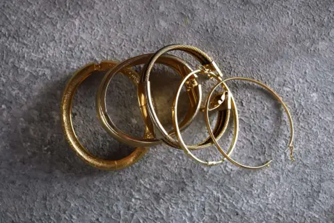 Un grupo de pulseras y aros dorados sobre fondo gris. Aunque el oro no se oxida, estas piezas lucen un brillo que evoca el paso del tiempo.