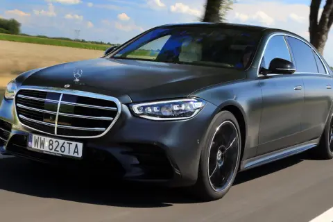 Elegancki, ciemnoszary Mercedes S580e sunie po drodze wśród zielonych pól i drzew.