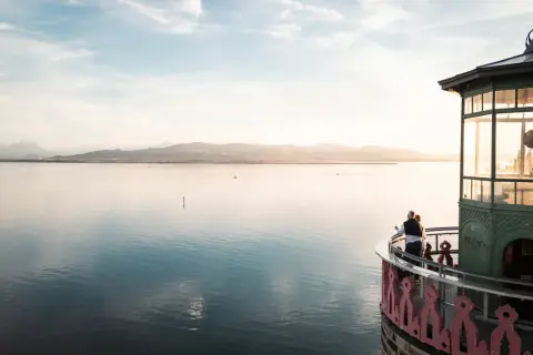 Ein Paar genießt den Sonnenuntergang am Leuchtturm Lindau. Der Bodensee glänzt, Berge im Hintergrund.