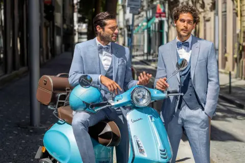 Zwei stilvolle Männer in hellblauen Anzügen, perfekt für einen Hochzeit Gast, posieren auf einer türkisfarbenen Vespa.
