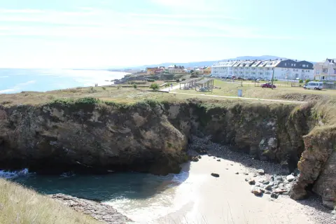 Acantilados rocosos y una pequeña cala de arena en la playa de San Bartolo Barreiros, con edificios residenciales al fondo.