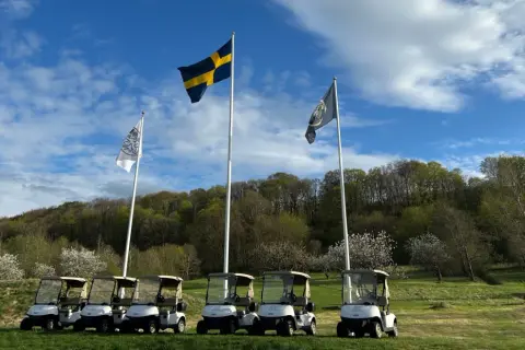 Golfbilar redo för spel vid äppelgården i Båstad. Svenska flaggan vajar stolt i bakgrunden.