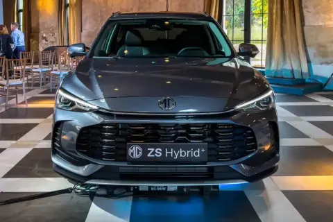 Szary MG ZS Hybrid+ prezentuje się elegancko na tle wnętrza z zasłonami i krzesłami.