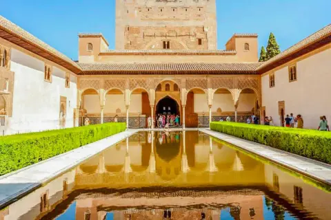 El Patio de los Leones en la Alhambra, con su estanque reflejando la arquitectura y un grupo de visitantes admirando la belleza.