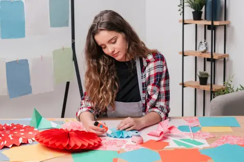 Chica creando manualidades para niños de 12 a 15 años. Pliegues de papel, abanicos y adornos coloridos sobre la mesa.