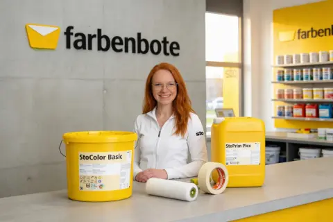 Frau mit roten Haaren lächelt, neben Farbeimer, Rolle und Klebeband. Alles, was man zum Streichen braucht, ist hier zu sehen.