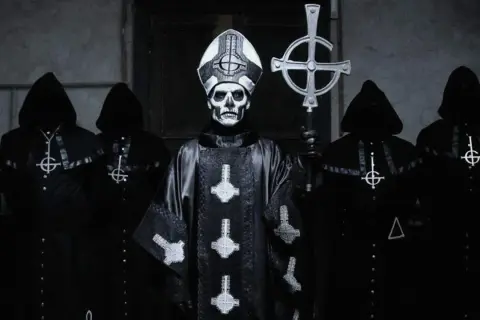Geister-Frontmann mit Totenkopf-Make-up und Kapuzengewänder, die an beliebte Metal-Bands erinnern.