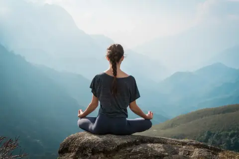 Frau in Hatha Yoga Pose auf einem Felsen sitzend, mit Blick auf eine neblige Berglandschaft.