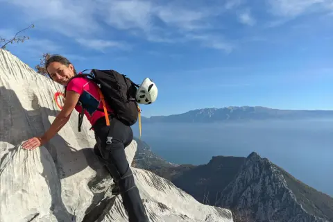 Donna sorridente su roccia con zaino e casco, vista lago e montagne. Un facile sentiero sul monte Pizzocolo.