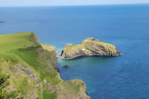 Falaises verdoyantes de la Causeway Coast, avec une île rocheuse reliée par un pont suspendu. L'eau bleue claire invite à la découverte.