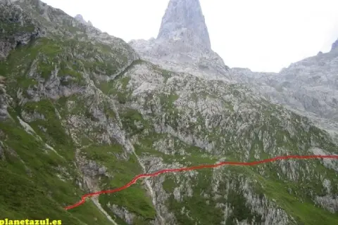 Sendero marcado en rojo asciende por la ladera verde hacia el imponente Sotres Urriellu, una cumbre rocosa en los Picos de Europa.