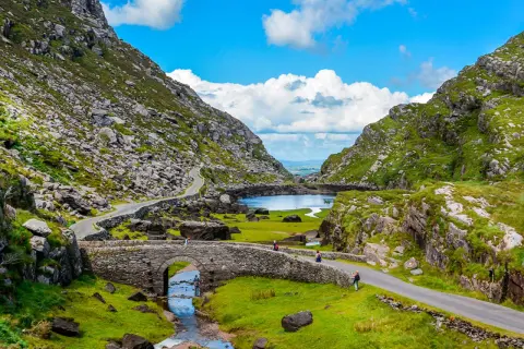 Eine malerische **irland landschaft** mit einem steinernen Bogenbrücken über einem Bach, der in einen See mündet, umgeben von grünen Hügeln und felsigen Bergen.