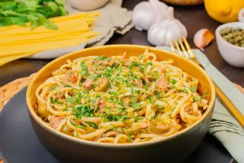 Eine Schüssel mit Thunfisch Pasta, garniert mit Petersilie und Zitronenschale. Daneben liegen Spaghetti, Knoblauch und Kapern.