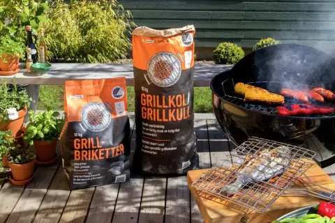 Två säckar med grillkol eller briketter står redo för grillkvällen. Majskolvar, korvar och paprika ryker på grillen.