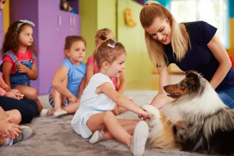 Uśmiechnięta dziewczynka głaszcze psa terapeutycznego, siedząc w grupie dzieci.