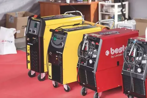 Trzy spawarki: czarna, żółta i czerwona, gotowe do pracy. Każda z nich ma uchwyt i kable, a czerwona posiada napis "best by Lincoln Electric".