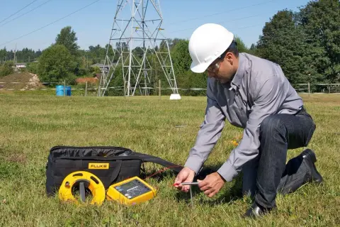 Technicien en plein air vérifiant la valeur prise de terre d'une maison, près d'un pylône électrique.