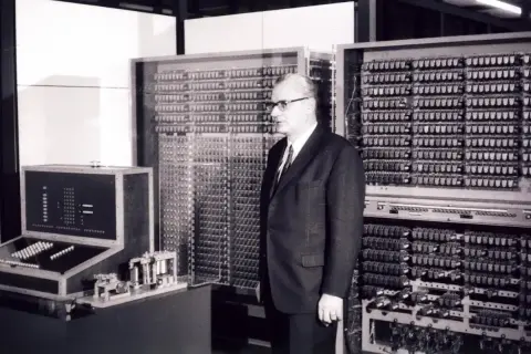 Mężczyzna w garniturze stoi obok ogromnej maszyny z mnóstwem przełączników i lamp. To prawdopodobnie pierwszy komputer na świecie, przełomowa technologia.