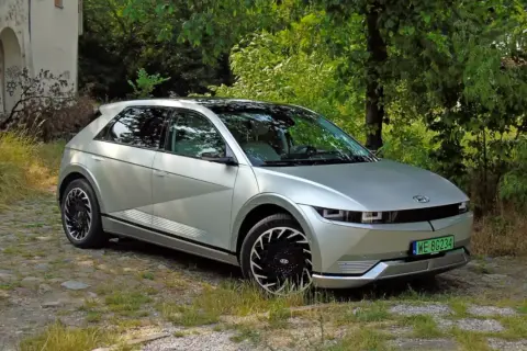 Srebrny Hyundai Ioniq 5 z czarnymi felgami stoi na kamienistej drodze, otoczony zielenią i starym budynkiem.