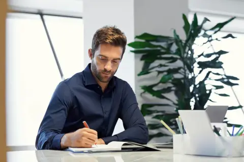 Hombre concentrado escribiendo en un diario en su escritorio, rodeado de plantas y un portátil.