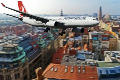 Samolot Turkish Airlines przelatuje nad miastem, jakby zmierzał w kierunku **Zakaukazia**. W tle widać zabytkowe budynki z czerwonymi dachami.