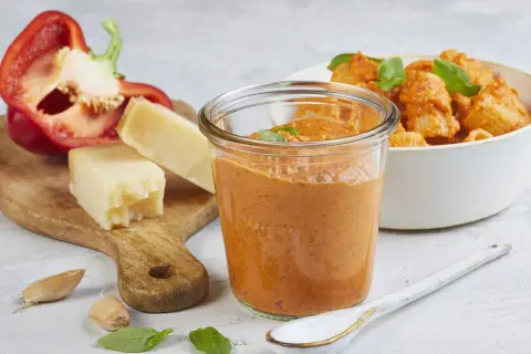 Ein Glas mit cremigem Paprika Pesto, daneben ein halber roter Paprika, Parmesan und Knoblauch. Im Hintergrund eine Schüssel Pasta mit dem Pesto.