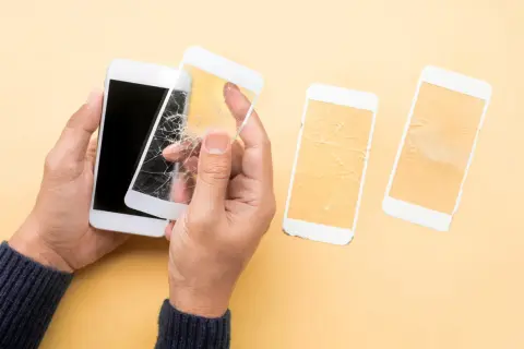 Ręce trzymają biały telefon z pękniętym szkłem hartowanym 5d. Obok leżą dwa inne telefony z rozbitymi ekranami.