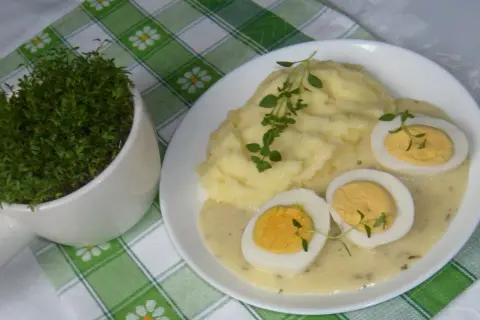Pyszne ziemniaki puree z jajkami w sosie chrzanowym na ciepło, udekorowane gałązkami ziół. Obok doniczka ze świeżą rzeżuchą.