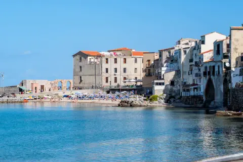 Edificios antiguos y una playa animada en Cefalú, Italia, con el mar azul brillante.