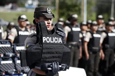 Agente de policía sostiene chaleco antibalas de material kevlar. Detrás, otros agentes uniformados.