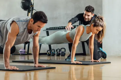 Trening funkcjonalny w grupie: mężczyzna i kobieta wykonują ćwiczenia w pozycji deski, a trener koryguje ich postawę.